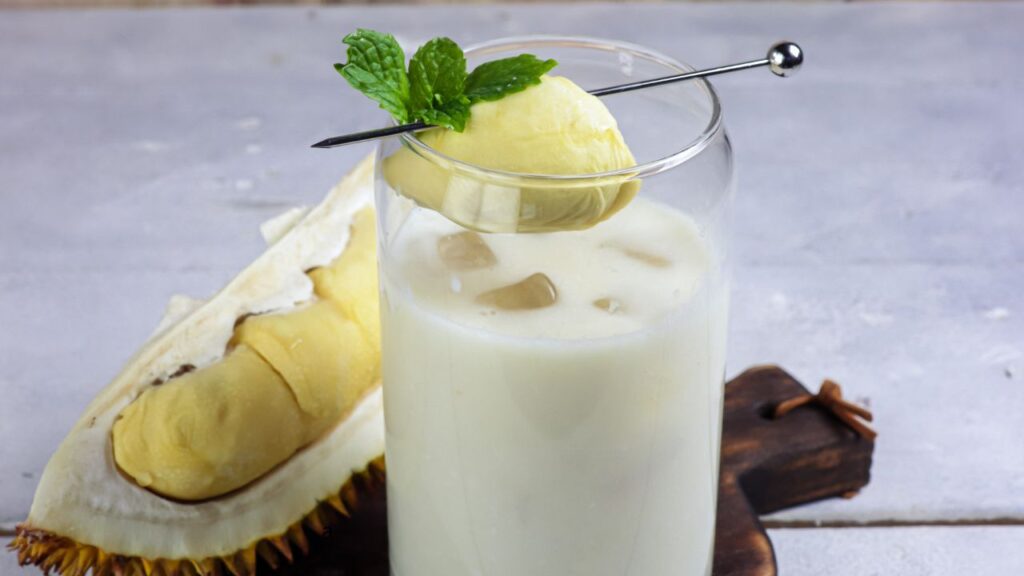 Smoothie durian creamy disajikan dalam gelas transparan, dilengkapi topping kacang cincang dan saus cokelat